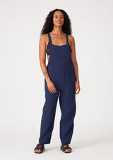 [Color: Royal] A front facing image of a brunette model wearing a soft cotton gauze bohemian lounge jumpsuit in royal blue. With the silhouette of overalls, featuring tank top straps that attach with a button closure, two side pockets, a wide leg, and a racer back.