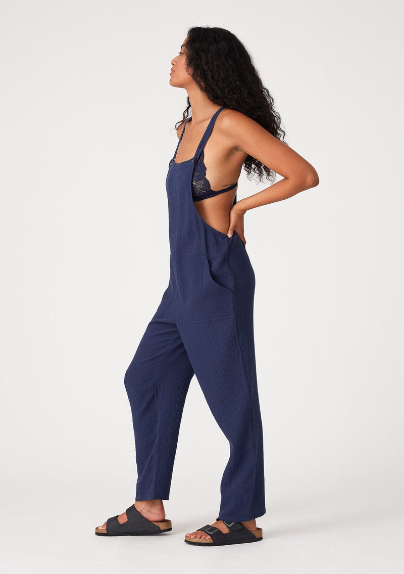 [Color: Royal] A side facing image of a brunette model wearing a soft cotton gauze bohemian lounge jumpsuit in royal blue. With the silhouette of overalls, featuring tank top straps that attach with a button closure, two side pockets, a wide leg, and a racer back.