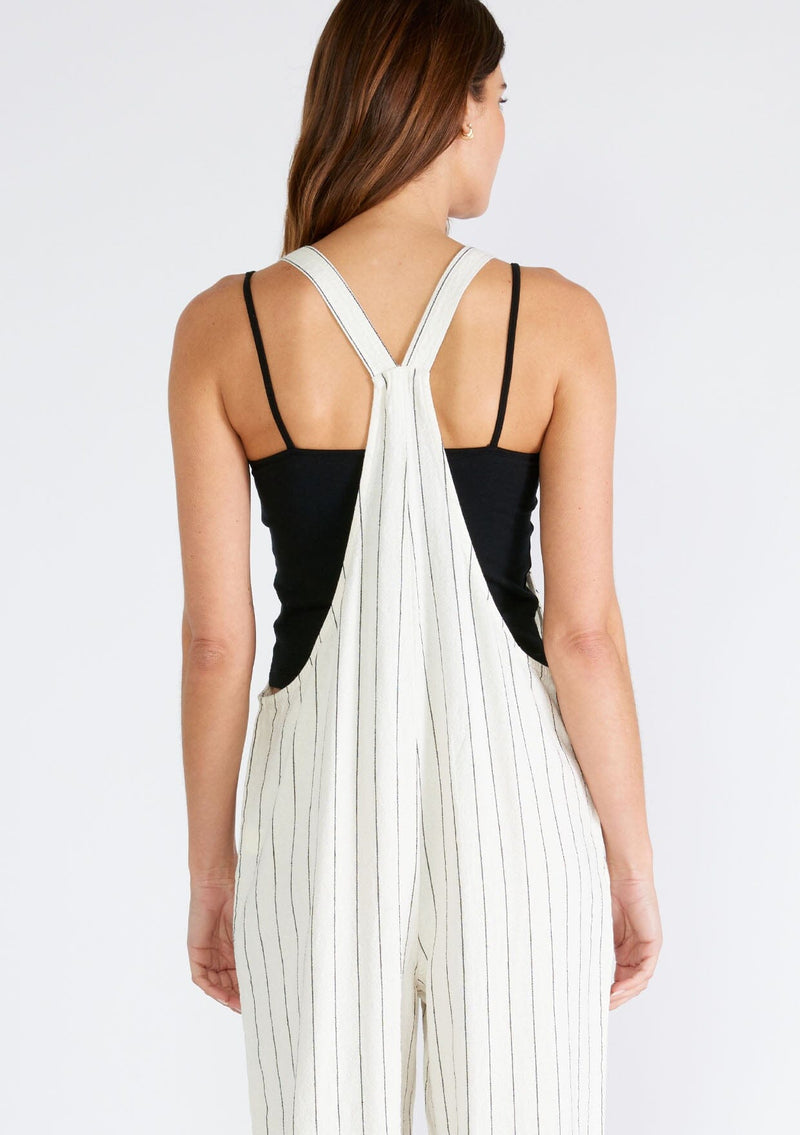 [Color: Natural/Black] A close up back facing image of a brunette model wearing a cool sleeveless jumpsuit crafted from a linen blend, in an ivory and black stripe. With a long straight leg, a racerback, tank top straps that button at the front, and side pockets. 
