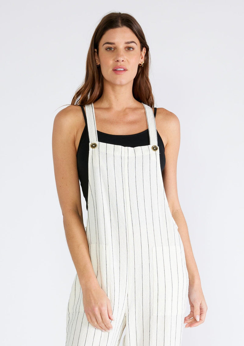 [Color: Natural/Black] A close up front facing image of a brunette model wearing a cool sleeveless jumpsuit crafted from a linen blend, in an ivory and black stripe. With a long straight leg, a racerback, tank top straps that button at the front, and side pockets. 