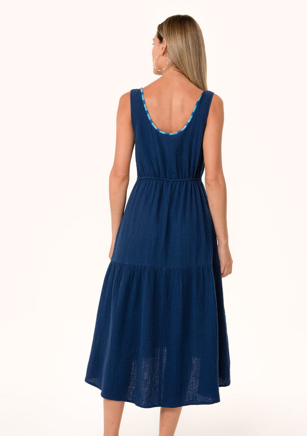 [Color: Navy] A back facing image of a blonde model wearing a navy blue cotton sleeveless mid length dress. With a scooped neckline, a tiered flowy skirt, a drawstring tassel tie waist, and contrast thread details. 