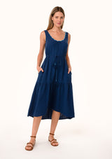 [Color: Navy] A front facing image of a blonde model wearing a navy blue cotton sleeveless mid length dress. With a scooped neckline, a tiered flowy skirt, a drawstring tassel tie waist, and contrast thread details. 