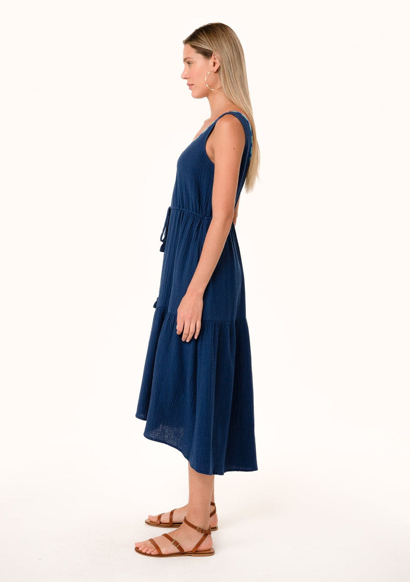 [Color: Navy] A side facing image of a blonde model wearing a navy blue cotton sleeveless mid length dress. With a scooped neckline, a tiered flowy skirt, a drawstring tassel tie waist, and contrast thread details. 