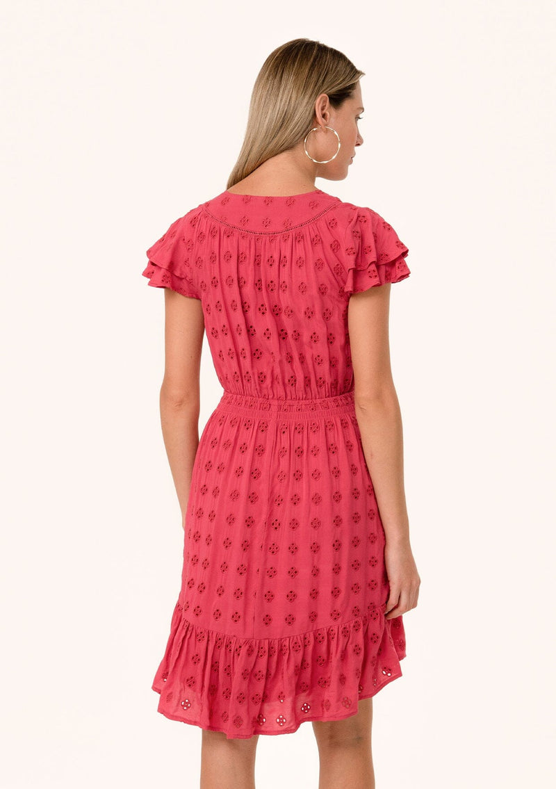 [Color: Fuchsia] A back facing image of a blonde model wearing a pink bohemian summer mini dress in embroidered eyelet. With short flutter sleeves, a smocked elastic waist, a v neckline, a tie waist accent, a tiered high low mini skirt, and lattice trim.