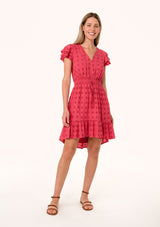 [Color: Fuchsia] A front facing image of a blonde model wearing a pink bohemian summer mini dress in embroidered eyelet. With short flutter sleeves, a smocked elastic waist, a v neckline, a tie waist accent, a tiered high low mini skirt, and lattice trim.