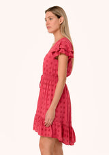 [Color: Fuchsia] A side facing image of a blonde model wearing a pink bohemian summer mini dress in embroidered eyelet. With short flutter sleeves, a smocked elastic waist, a v neckline, a tie waist accent, a tiered high low mini skirt, and lattice trim.