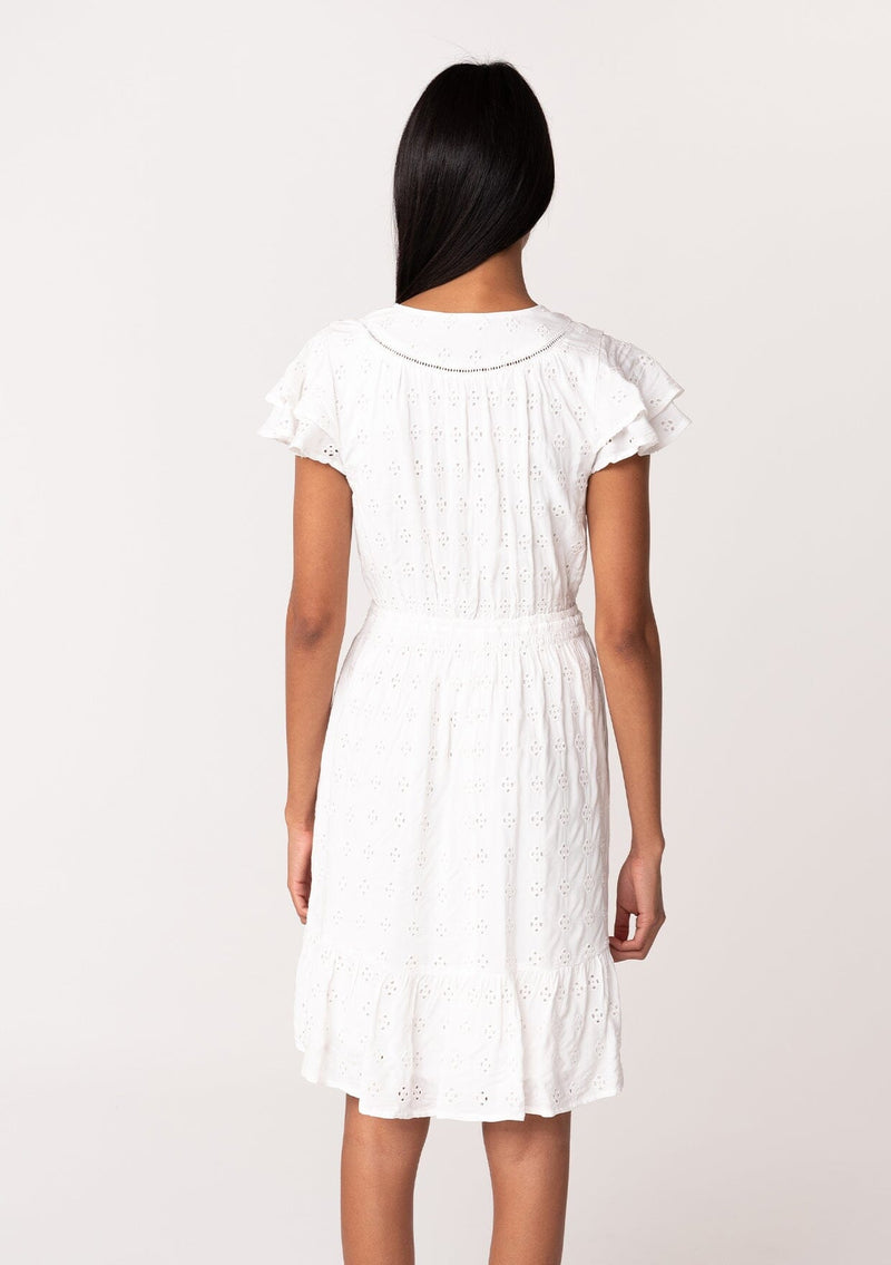 [Color: White] A back facing image of a brunette model wearing a white bohemian summer mini dress in embroidered eyelet. With short flutter sleeves, a smocked elastic waist, a v neckline, a tie waist accent, a tiered high low mini skirt, and lattice trim. 