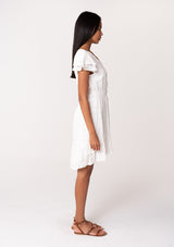 [Color: White] A side facing image of a brunette model wearing a white bohemian summer mini dress in embroidered eyelet. With short flutter sleeves, a smocked elastic waist, a v neckline, a tie waist accent, a tiered high low mini skirt, and lattice trim. 