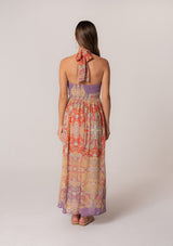 [Color: Red/Purple] A full body back facing image of a brunette model wearing a bohemian halter maxi dress in a multi colored red and purple floral print. With a tie neckline, a slim fit bodice with a smocked elastic back, and a long flowy skirt.