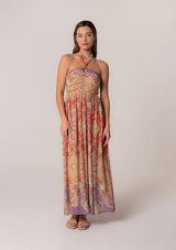 [Color: Red/Purple] A full body front facing image of a brunette model wearing a bohemian halter maxi dress in a multi colored red and purple floral print. With a tie neckline, a slim fit bodice with a smocked elastic back, and a long flowy skirt.