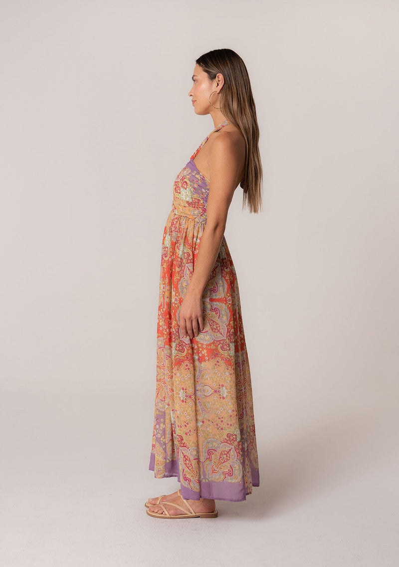 [Color: Red/Purple] A full body side facing image of a brunette model wearing a bohemian halter maxi dress in a multi colored red and purple floral print. With a tie neckline, a slim fit bodice with a smocked elastic back, and a long flowy skirt.