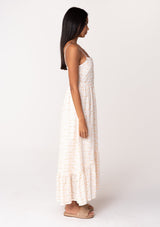 [Color: White/Apricot] A side facing image of a brunette model wearing a cotton bohemian maxi dress in a white and pink embroidered floral design. With adjustable spaghetti straps, a straight neckline, a button front, a half smocked slim fit bodice at the back, and a flowy long tiered skirt. 
