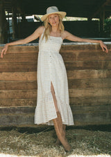 [Color: White/Apricot] A front facing image of a blonde model standing outside wearing a cotton bohemian maxi dress in a white and pink embroidered floral design. With adjustable spaghetti straps, a straight neckline, a button front, a half smocked slim fit bodice at the back, and a flowy long tiered skirt.