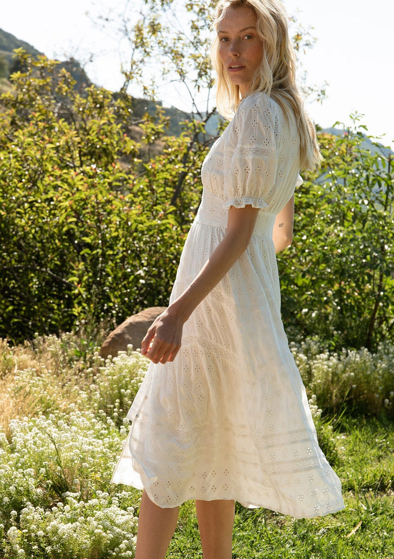 [Color: White] A side facing image of a blonde model standing in a field wearing a white bohemian mid length dress in embroidered eyelet. With a flowy lace trimmed tiered skirt, a button front, short puff sleeves, and a lace trimmed v neckline.