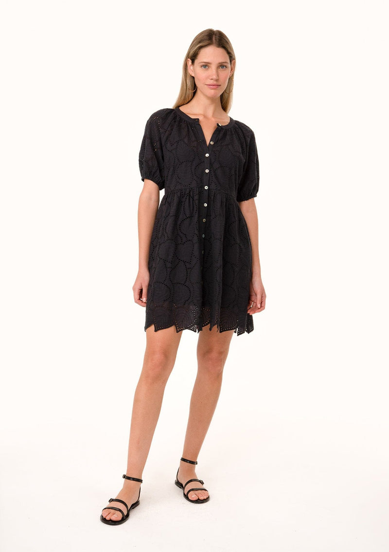 [Color: Black] A front facing image of a blonde model wearing a black summer mini dress in embroidered eyelet. With short puff sleeves, a round neckline, a button front, and a relaxed loose fit.