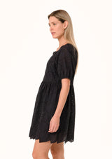 [Color: Black] A side facing image of a blonde model wearing a black summer mini dress in embroidered eyelet. With short puff sleeves, a round neckline, a button front, and a relaxed loose fit.