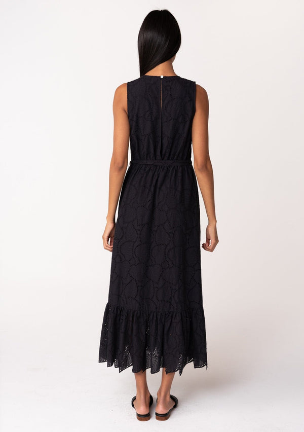 [Color: Black] A back facing image of a brunette model wearing a sleeveless black bohemian summer midi dress in embroidered eyelet. With a v neckline, a flowy tiered skirt, a self tie waist belt, and a back keyhole with button closure. 