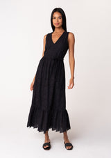 [Color: Black] A front facing image of a brunette model wearing a sleeveless black bohemian summer midi dress in embroidered eyelet. With a v neckline, a flowy tiered skirt, a self tie waist belt, and a back keyhole with button closure. 