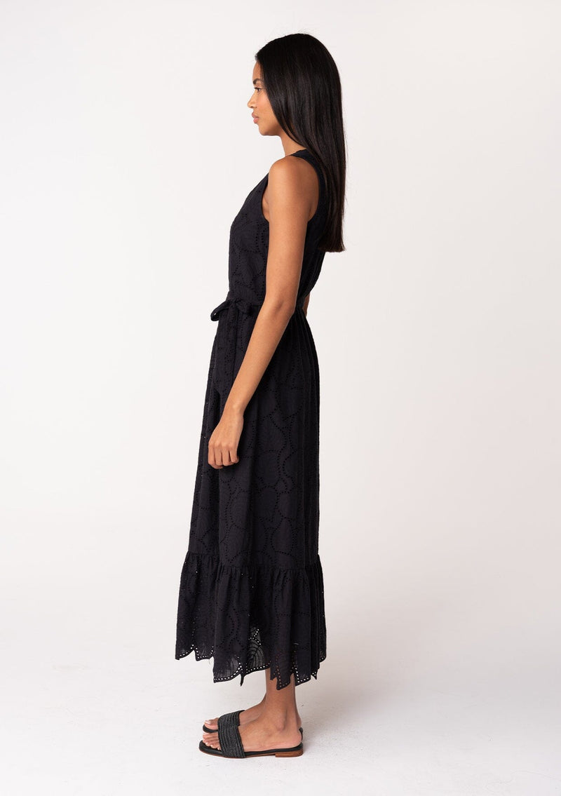 [Color: Black] A side facing image of a brunette model wearing a sleeveless black bohemian summer midi dress in embroidered eyelet. With a v neckline, a flowy tiered skirt, a self tie waist belt, and a back keyhole with button closure. 