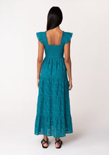 [Color: Teal] A back facing image of a brunette model wearing a bohemian cotton maxi dress in a teal embroidered eyelet. With a slim fit smocked bodice, a square neckline, short flutter sleeves, and a flowy long tiered skirt. 