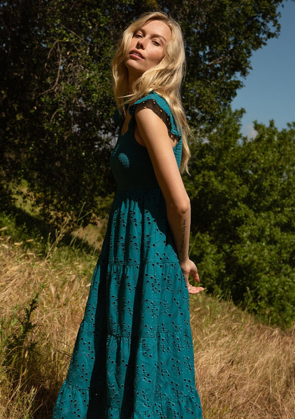 [Color: Teal] A close up side facing image of a blonde model standing in a field wearing a bohemian cotton maxi dress in a teal embroidered eyelet. With a slim fit smocked bodice, a square neckline, short flutter sleeves, and a flowy long tiered skirt.