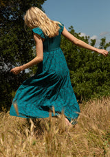 [Color: Teal] A back facing image of a blonde model standing in a field wearing a bohemian cotton maxi dress in a teal embroidered eyelet. With a slim fit smocked bodice, a square neckline, short flutter sleeves, and a flowy long tiered skirt.