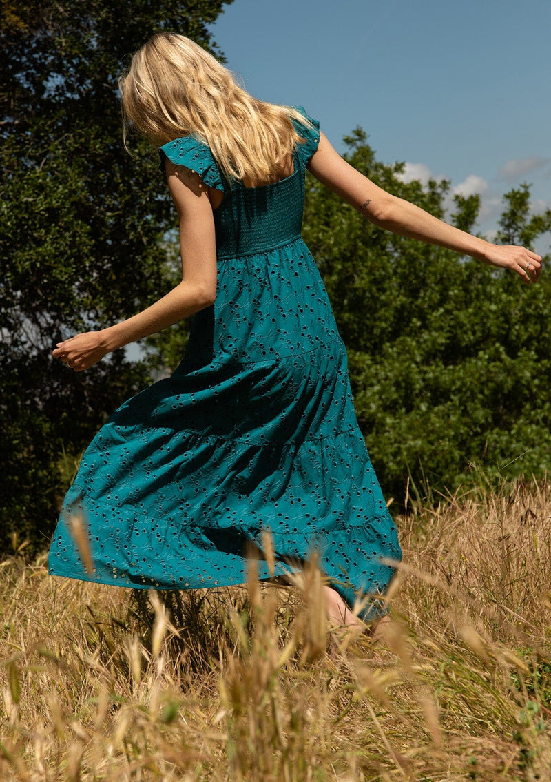 [Color: Teal] A back facing image of a blonde model standing in a field wearing a bohemian cotton maxi dress in a teal embroidered eyelet. With a slim fit smocked bodice, a square neckline, short flutter sleeves, and a flowy long tiered skirt.
