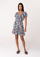 [Color: Navy/Natural] A full body front facing image of a brunette model wearing a navy blue floral print summer mini dress. With short flutter sleeves, a split v neckline with tassel ties, an elastic waist, a self covered button front top, and a tiered mini skirt. 