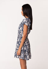 [Color: Navy/Natural] A side facing image of a brunette model wearing a navy blue floral print summer mini dress. With short flutter sleeves, a split v neckline with tassel ties, an elastic waist, a self covered button front top, and a tiered mini skirt. 