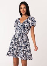 [Color: Navy/Natural] A front facing image of a brunette model wearing a navy blue floral print summer mini dress. With short flutter sleeves, a split v neckline with tassel ties, an elastic waist, a self covered button front top, and a tiered mini skirt. 