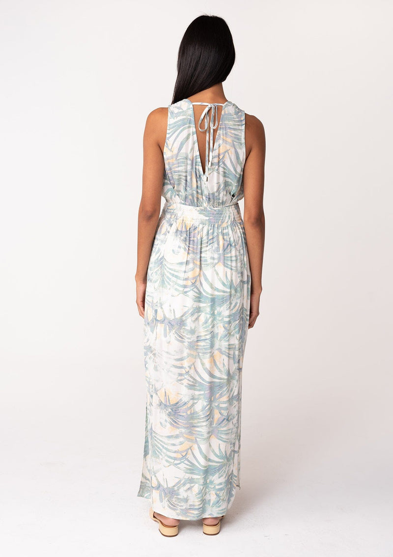 [Color: Natural/Seafoam] A back facing image of a brunette model wearing a sleeveless bohemian summer maxi dress in a green palm leaf print. With a smocked elastic waist, a v neckline, a flowy long skirt with side slits, and an open back with tie closure. 