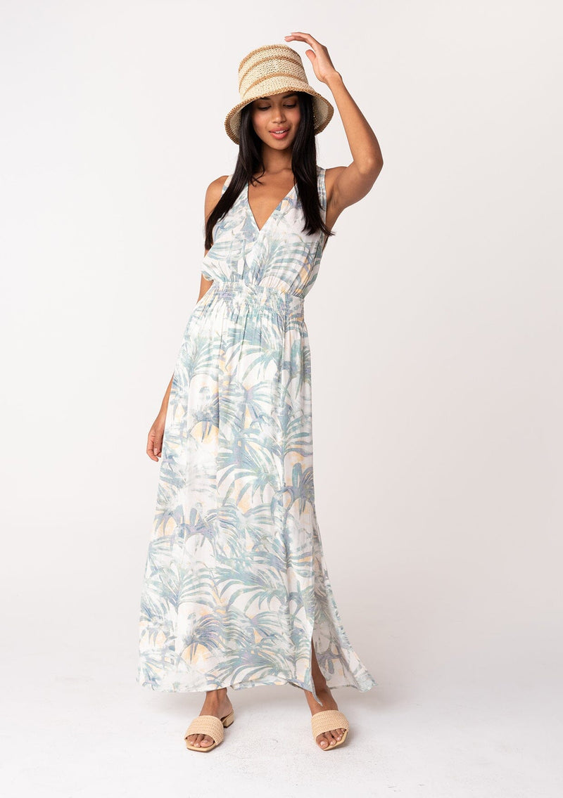 [Color: Natural/Seafoam] A front facing image of a brunette model wearing a sleeveless bohemian summer maxi dress in a green palm leaf print. With a smocked elastic waist, a v neckline, a flowy long skirt with side slits, and an open back with tie closure. 