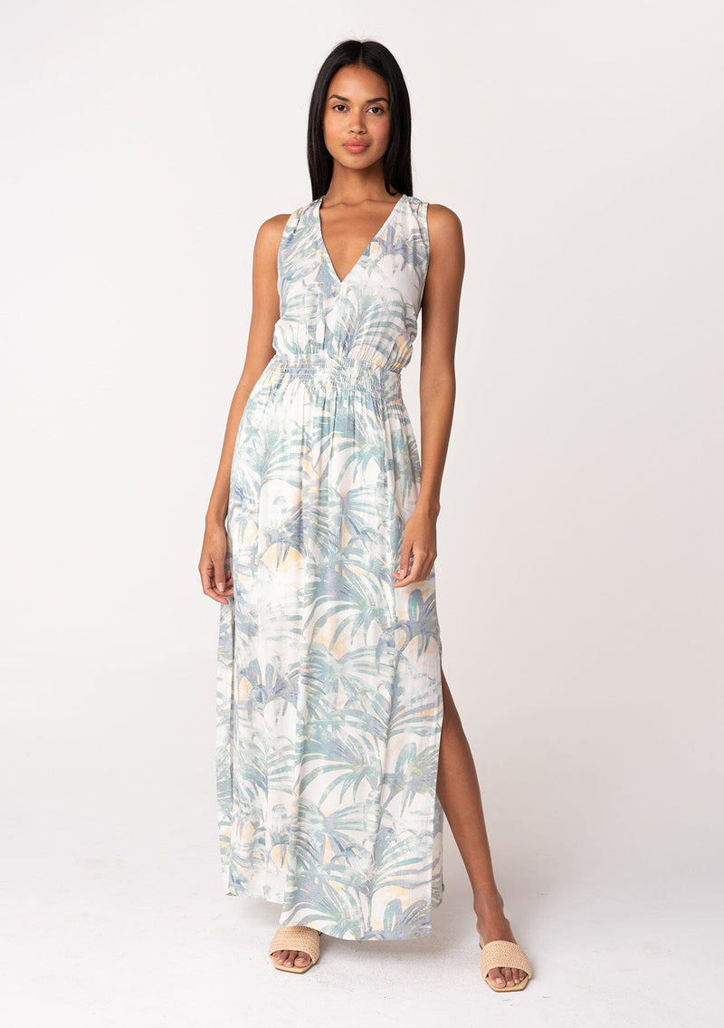 [Color: Natural/Seafoam] A full body front facing image of a brunette model wearing a sleeveless bohemian summer maxi dress in a green palm leaf print. With a smocked elastic waist, a v neckline, a flowy long skirt with side slits, and an open back with tie closure. 