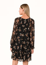 [Color: Black/Peach] A back facing image of a blonde model wearing a pretty bohemian chiffon mini dress in a black and peach floral print. With sheer long sleeves, ruffled elastic wrist cuffs, a tiered skirt, a v neckline with tassel ties, and a decorative self covered button front. 