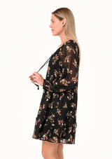 [Color: Black/Peach] A side facing image of a blonde model wearing a pretty bohemian chiffon mini dress in a black and peach floral print. With sheer long sleeves, ruffled elastic wrist cuffs, a tiered skirt, a v neckline with tassel ties, and a decorative self covered button front. 