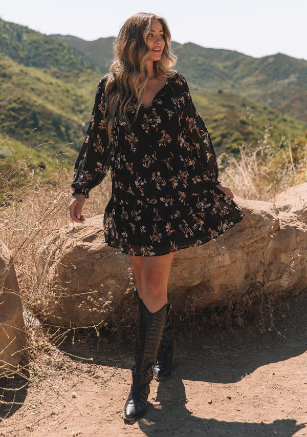 [Color: Black/Peach] A front facing image of a blonde model standing outside wearing a pretty bohemian chiffon mini dress in a black and peach floral print. With sheer long sleeves, ruffled elastic wrist cuffs, a tiered skirt, a v neckline with tassel ties, and a decorative self covered button front.