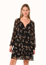 [Color: Black/Peach] A front facing image of a blonde model wearing a pretty bohemian chiffon mini dress in a black and peach floral print. With sheer long sleeves, ruffled elastic wrist cuffs, a tiered skirt, a v neckline with tassel ties, and a decorative self covered button front. 