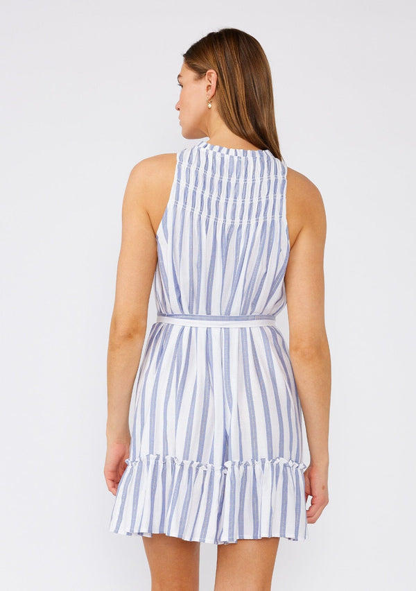 [Color: White/Blue] A back facing image of a brunette model wearing a sleeveless spring mini dress designed in a blue and white stripe and a gold metallic thread detail. With a flowy ruffled trimmed tiered skirt, a smocked yoke, a ruffled v neckline, and a self tie waist belt. 