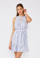 [Color: White/Blue] A front facing image of a brunette model wearing a sleeveless spring mini dress designed in a blue and white stripe and a gold metallic thread detail. With a flowy ruffled trimmed tiered skirt, a smocked yoke, a ruffled v neckline, and a self tie waist belt. 