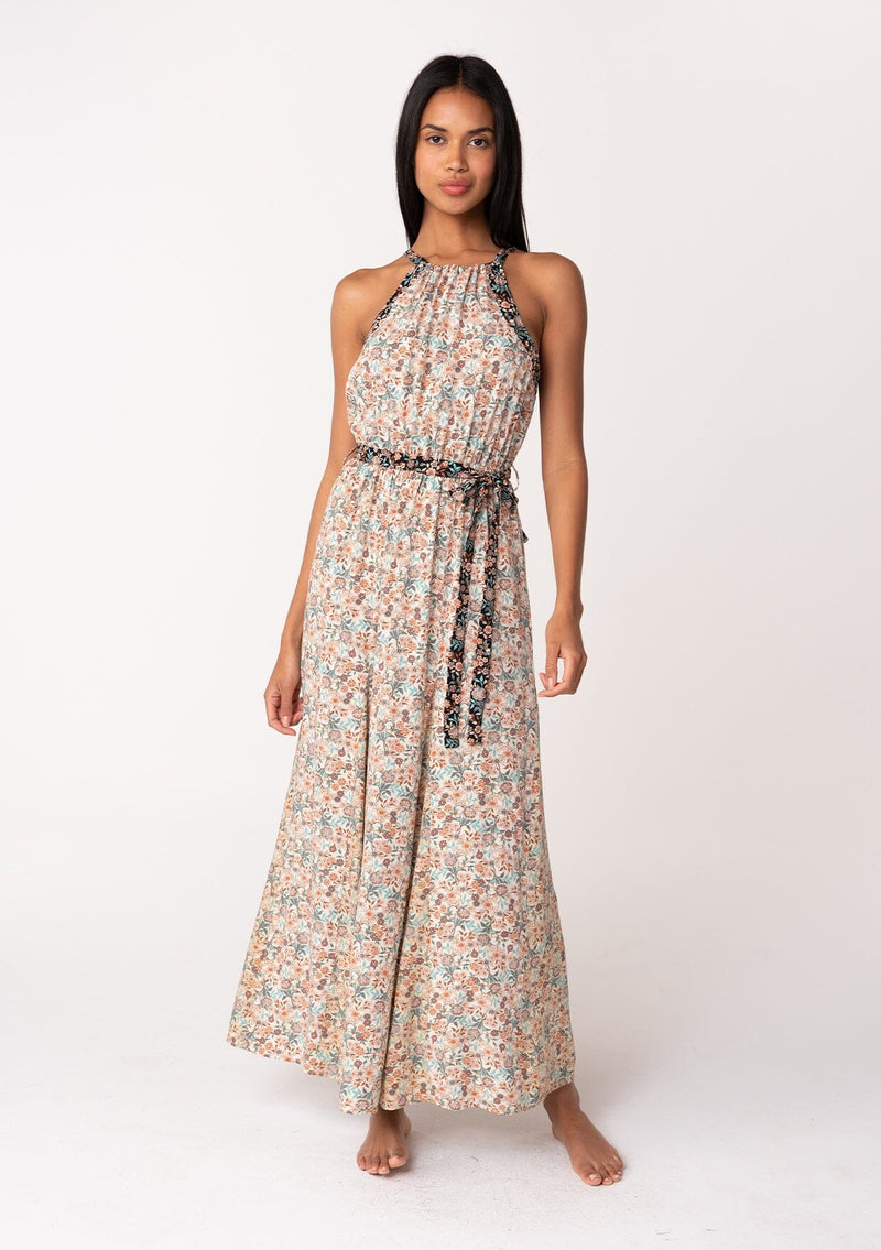 [Color: Taupe/Black] A front facing image of a brunette model wearing a fall floral sleeveless bohemian maxi dress in a taupe and black floral print. With a halter neckline, a drawstring tie neck, a back keyhole detail, a long flowy paneled skirt, an elastic waist, and a tie waist belt. 