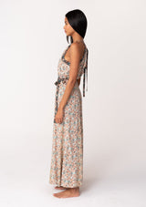 [Color: Taupe/Black] A side facing image of a brunette model wearing a fall floral sleeveless bohemian maxi dress in a taupe and black floral print. With a halter neckline, a drawstring tie neck, a back keyhole detail, a long flowy paneled skirt, an elastic waist, and a tie waist belt. 