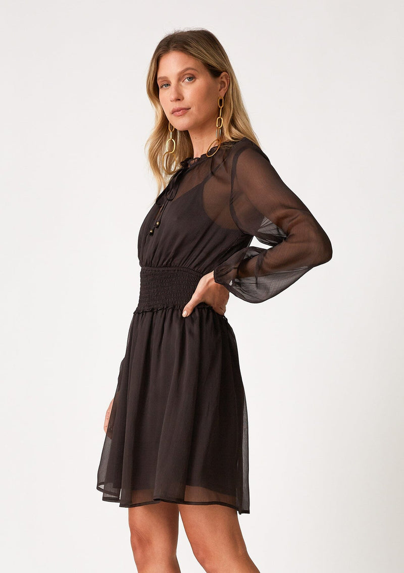 [Color: Chocolate] A side facing image of a blonde model wearing a bohemian fall mini dress in a sheer dark brown chiffon. With long sleeves, a smocked elastic waist, a ruffled neckline, and a split v neckline with ties.