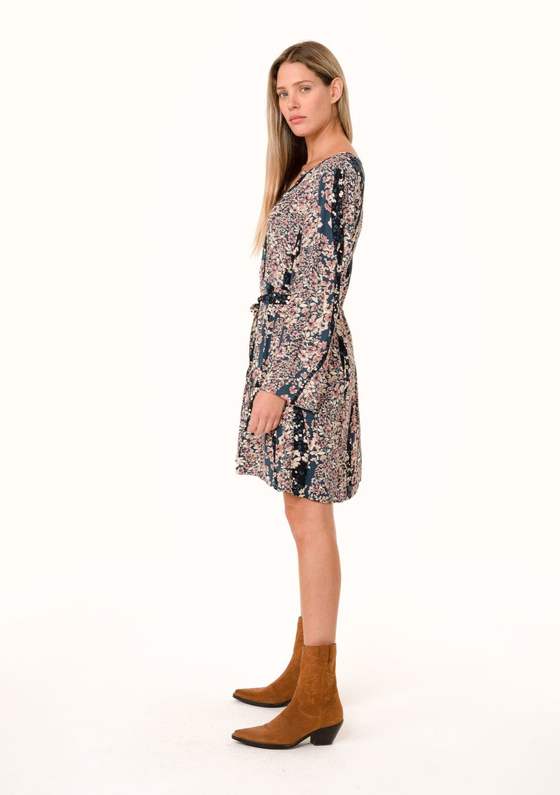 [Color: Dusty Rose/Navy] A side facing image of a blonde model wearing a bohemian fall mini dress in a blue and pink mixed floral print. With long sleeves, a split v neckline, a flowy paneled skirt, a button front, and an adjustable waist tie. 