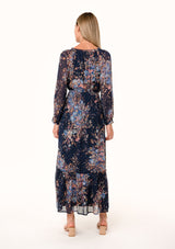 [Color: Navy/Dusty Blue] A back facing image of a blonde model wearing a bohemian fall maxi dress in a blue floral print. Designed in a metallic clip dot chiffon, with sheer long sleeves, a v neckline, a ruffle trimmed tiered skirt, a side slit, a smocked elastic waist, and a split v neckline with tassel ties. 