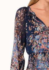 [Color: Navy/Dusty Blue] A close up front facing image of a blonde model wearing a bohemian fall maxi dress in a blue floral print. Designed in a metallic clip dot chiffon, with sheer long sleeves, a v neckline, a ruffle trimmed tiered skirt, a side slit, a smocked elastic waist, and a split v neckline with tassel ties. 