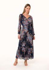 [Color: Navy/Dusty Blue] A front facing image of a blonde model wearing a bohemian fall maxi dress in a blue floral print. Designed in a metallic clip dot chiffon, with sheer long sleeves, a v neckline, a ruffle trimmed tiered skirt, a side slit, a smocked elastic waist, and a split v neckline with tassel ties. 