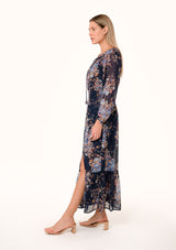 [Color: Navy/Dusty Blue] A side facing image of a blonde model wearing a bohemian fall maxi dress in a blue floral print. Designed in a metallic clip dot chiffon, with sheer long sleeves, a v neckline, a ruffle trimmed tiered skirt, a side slit, a smocked elastic waist, and a split v neckline with tassel ties. 