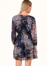[Color: Navy/Dusty Blue] A back facing image of a blonde model wearing a bohemian fall chiffon mini dress in a blue floral print. Designed with metallic clip dot details throughout, sheer long sleeves, a v neckline, an elastic waist, a decorative button front, and a double layered tiered skirt. 
