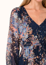 [Color: Navy/Dusty Blue] A close up front facing image of a blonde model wearing a bohemian fall chiffon mini dress in a blue floral print. Designed with metallic clip dot details throughout, sheer long sleeves, a v neckline, an elastic waist, a decorative button front, and a double layered tiered skirt. 
