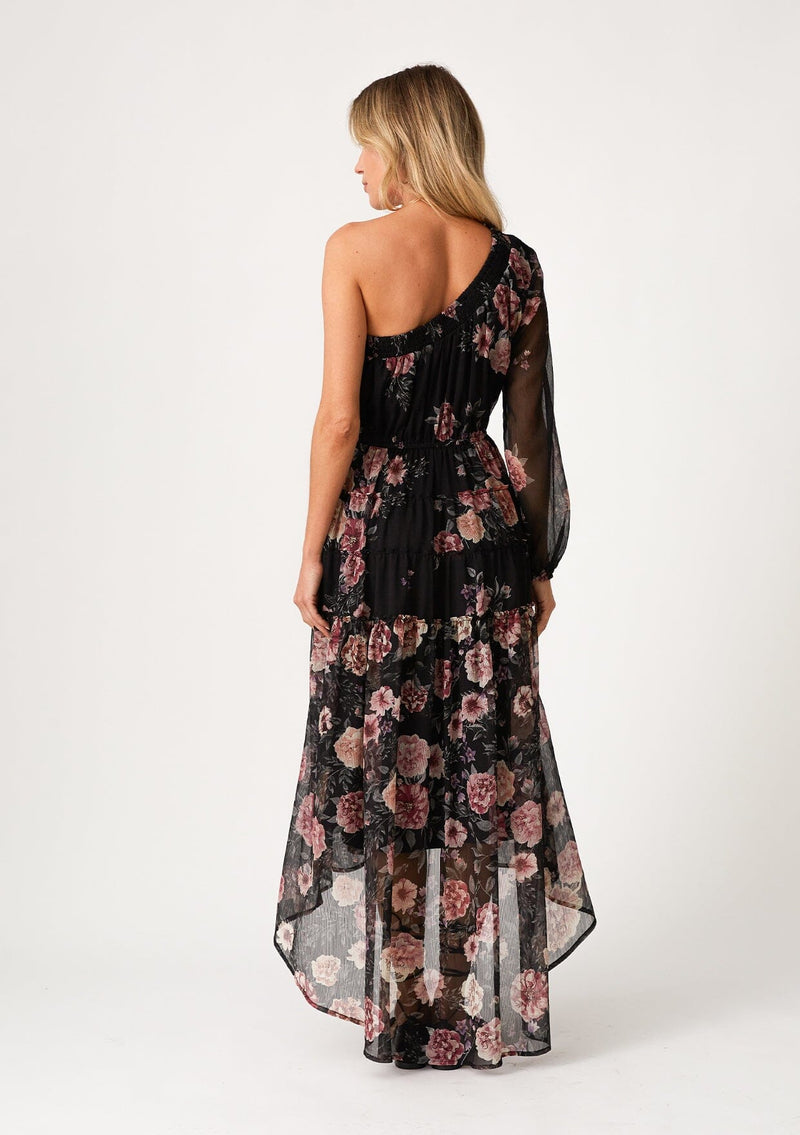 [Color: Black/Rose] A back facing image of a blonde model wearing a special occasion bohemian maxi dress in a black and pink floral print. Designed in chiffon, with a one shoulder detail, a long sleeve, a flowy ruffle trimmed tiered skirt, a high low hemline, an elastic waist, and a smocked elastic neckline. 